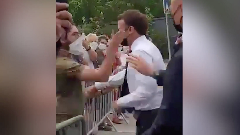 French President Emmanuel Macron, center, in white, was slapped Tuesday while on an official visit to a small town in southern France. The main suspect, left, in green, is in custody on charges of willful violence against a person holding public authority, according to French broadcaster BFM TV. Screenshot via Yahoo News.