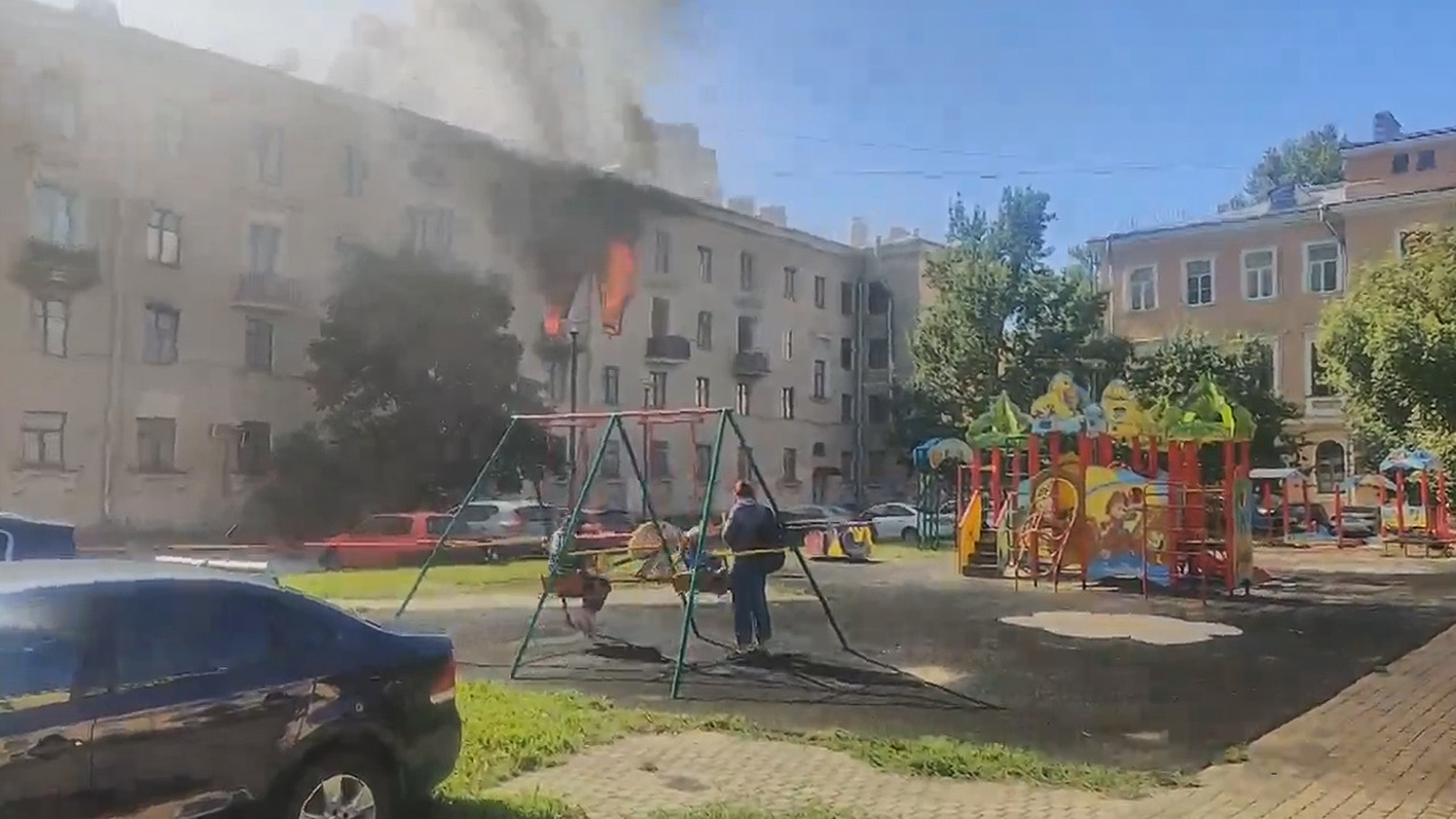 Two boys play on a swing set, apparently paying no heed to the fire in the nearby building. (Artyom Yakovlev/Zenger)